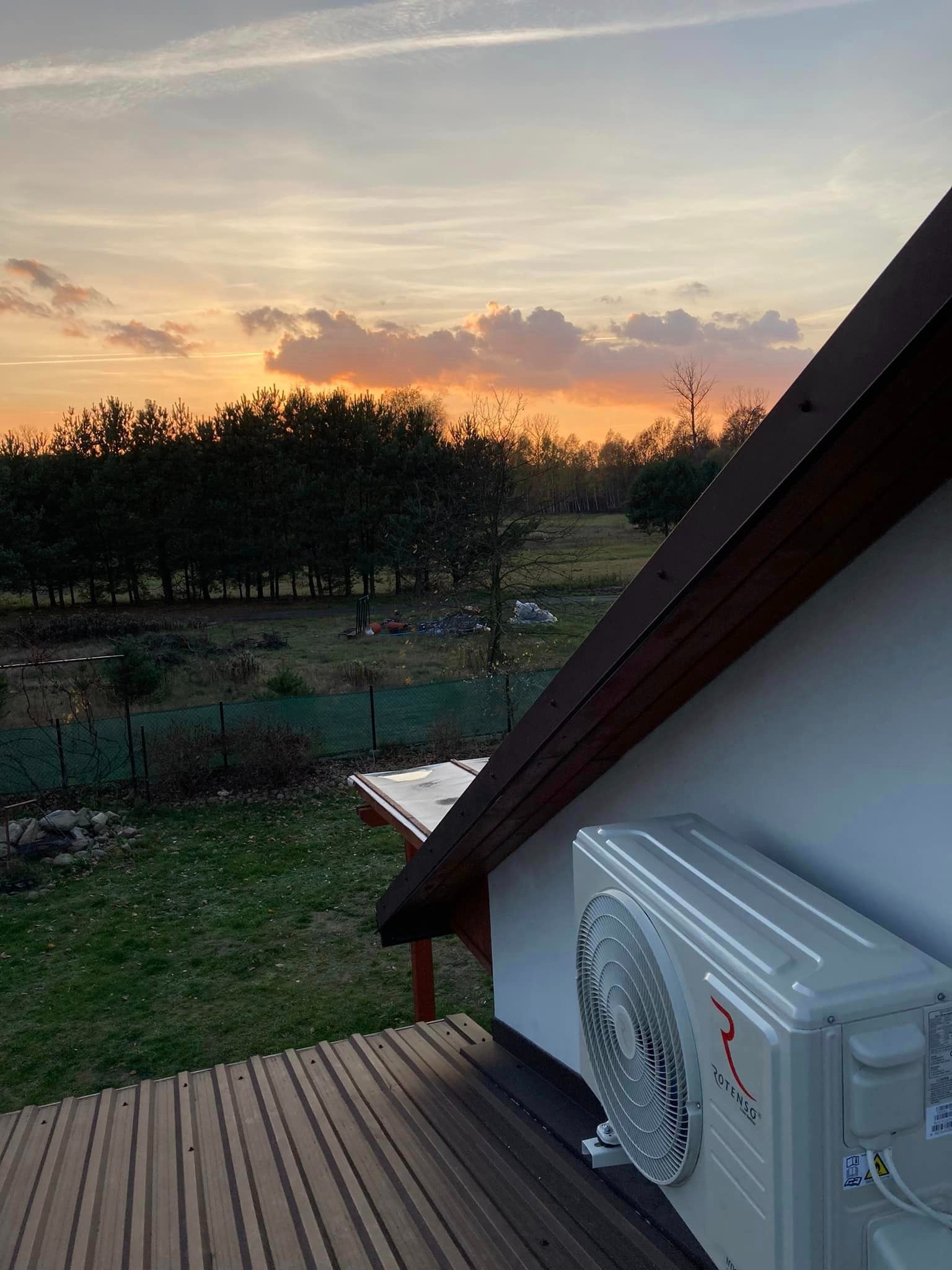 Zewnętrzna jednostka klimatyzacji marki Rotenso zamontowana na ścianie domu, widok z góry na tle zachodzącego słońca nad lasem.