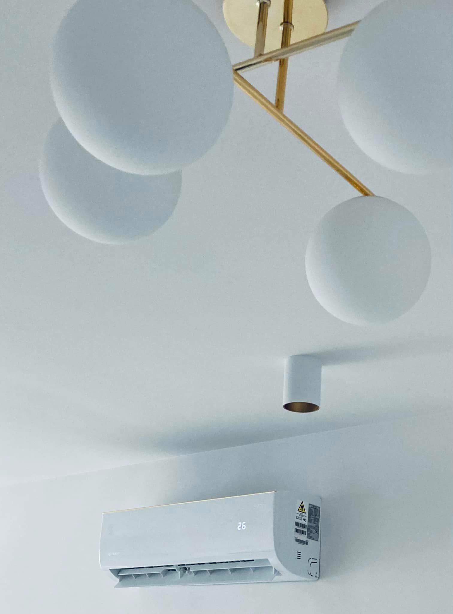 Nowoczesny, biały klimatyzator ścienny z wyświetlaczem temperatury (26 stopni Celsjusza) zamontowany pod sufitem z designerską lampą kulistą z elementami w kolorze złotym i minimalistycznym...