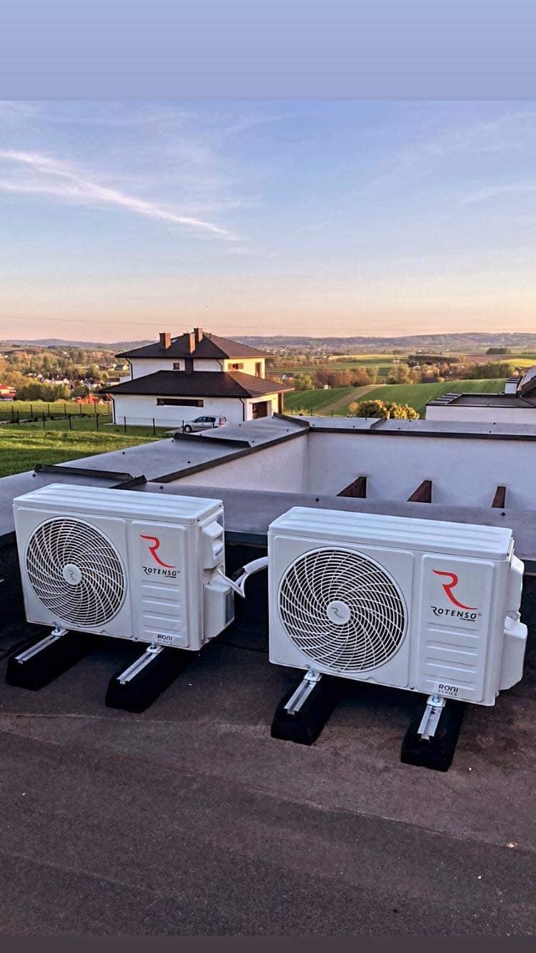 Dwa białe klimatyzatory marki Rotenso zamontowane na płaskim dachu domu jednorodzinnego z widokiem na malowniczy krajobraz wiejski o zachodzie słońca.