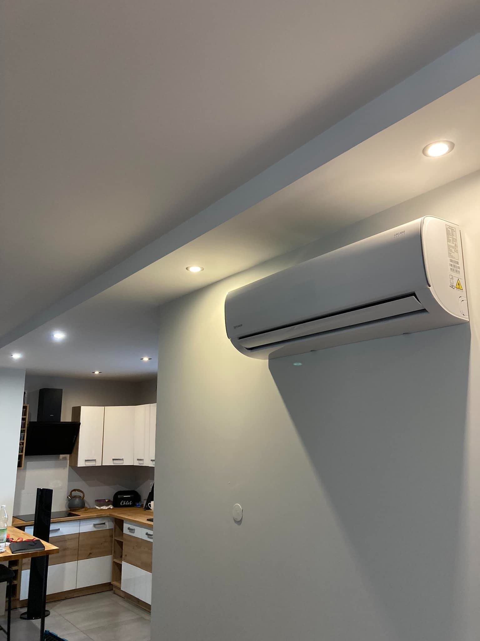 Biały klimatyzator ścienny zamontowany w nowoczesnym wnętrzu domu z widoczną kuchnią w tle, oświetlony punktowymi lampami sufitowymi.