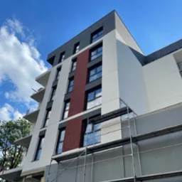 Wielopiętrowy budynek z częściowo otynkowaną elewacją, widoczne rusztowania i fragmenty ocieplenia, błękitne niebo w tle.