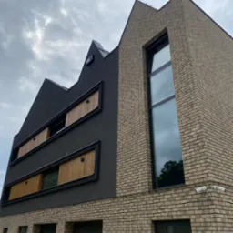 Nowoczesna elewacja budynku z cegły klinkierowej i szarego tynku, z drewnianymi elementami przesłon okiennych, widoczne kamery monitoringu i brama garażowa