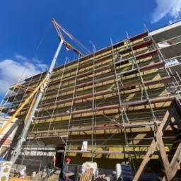 Budynek w trakcie ocieplania, widoczne rusztowania, dźwig i pracownicy w czerwonych czapkach, składowane materiały budowlane na dole, błękitne niebo w tle.