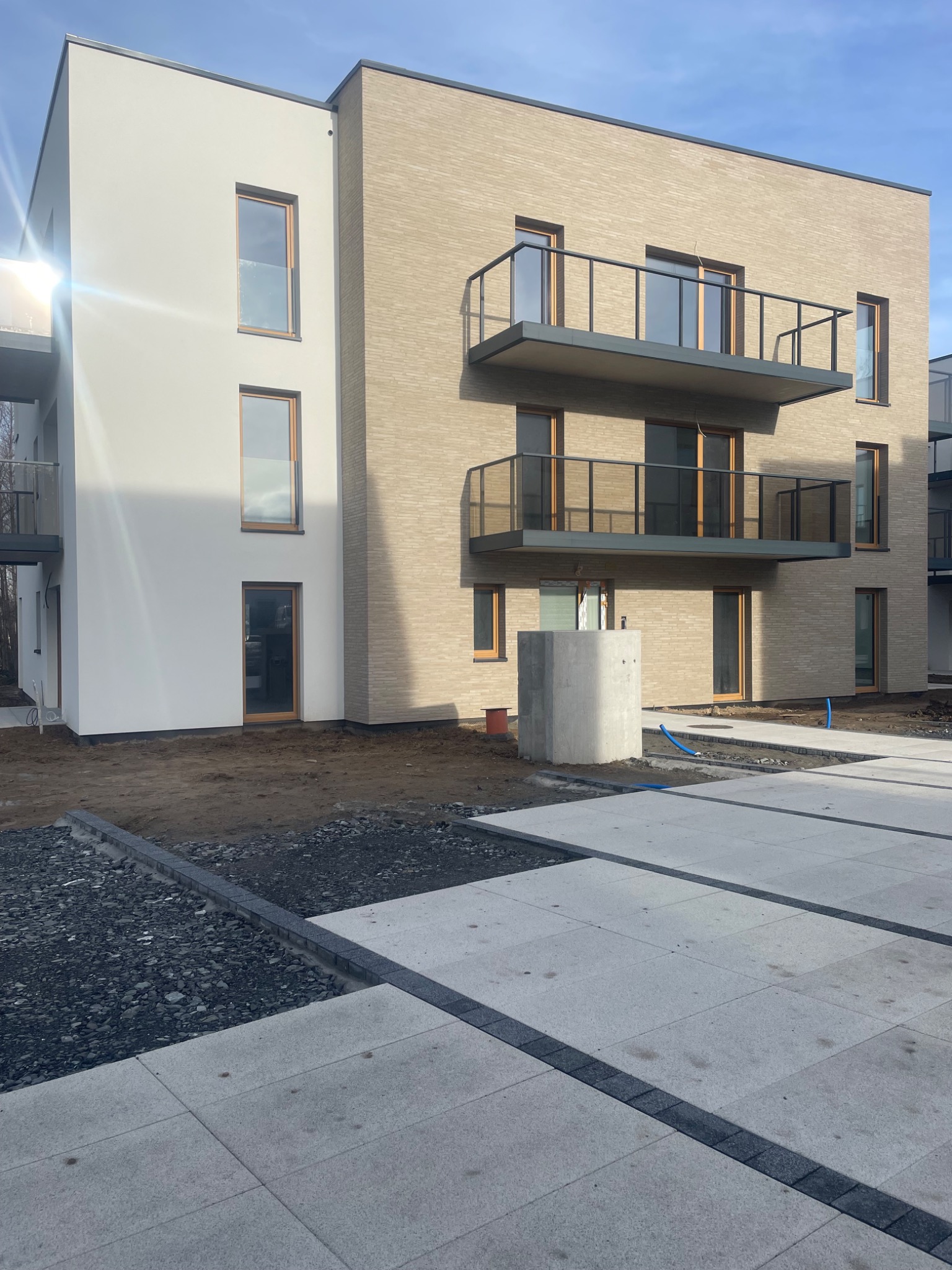 Nowoczesny budynek wielorodzinny z elewacją w dwóch odcieniach: białym i beżowym, z balkonami z szklanymi balustradami, widoczne prace wykończeniowe wokół budynku.