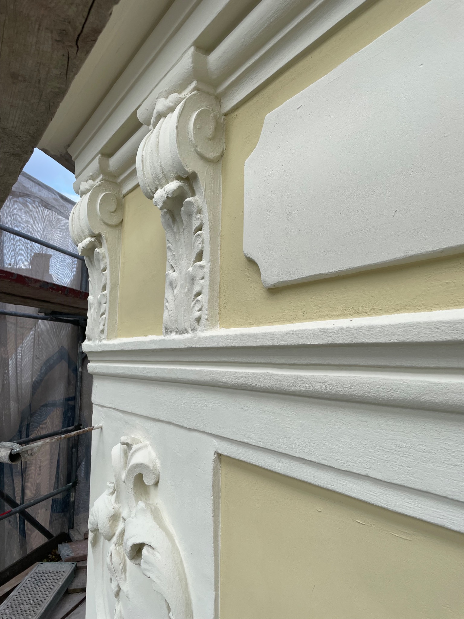 Fragment odnowionej elewacji z detalami sztukaterii w kolorze białym na jasnożółtym tle, widoczne rusztowanie z lewej strony.
