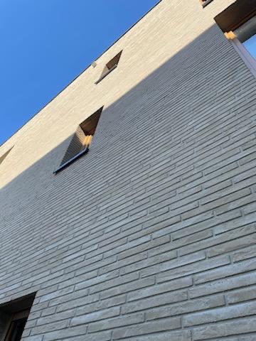 Nowoczesna elewacja budynku z jasnej cegły klinkierowej, widok z dołu na fasadę i okna o nietypowym kształcie pod błękitnym niebem.