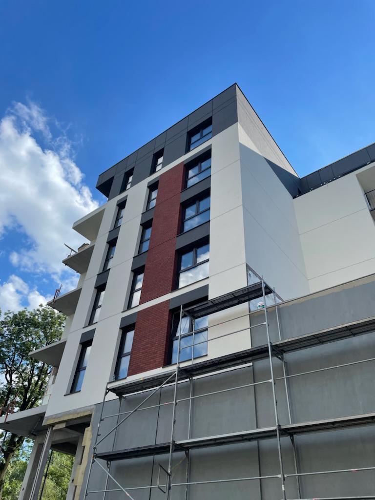 Wielopiętrowy budynek z częściowo otynkowaną elewacją, widoczne rusztowania i fragmenty ocieplenia, błękitne niebo w tle.