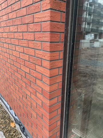 Fragment elewacji z czerwonej cegły klinkierowej, narożnik budynku i okno z czarną ramą, widoczne detale spoinowania i podłoże z kamykami.