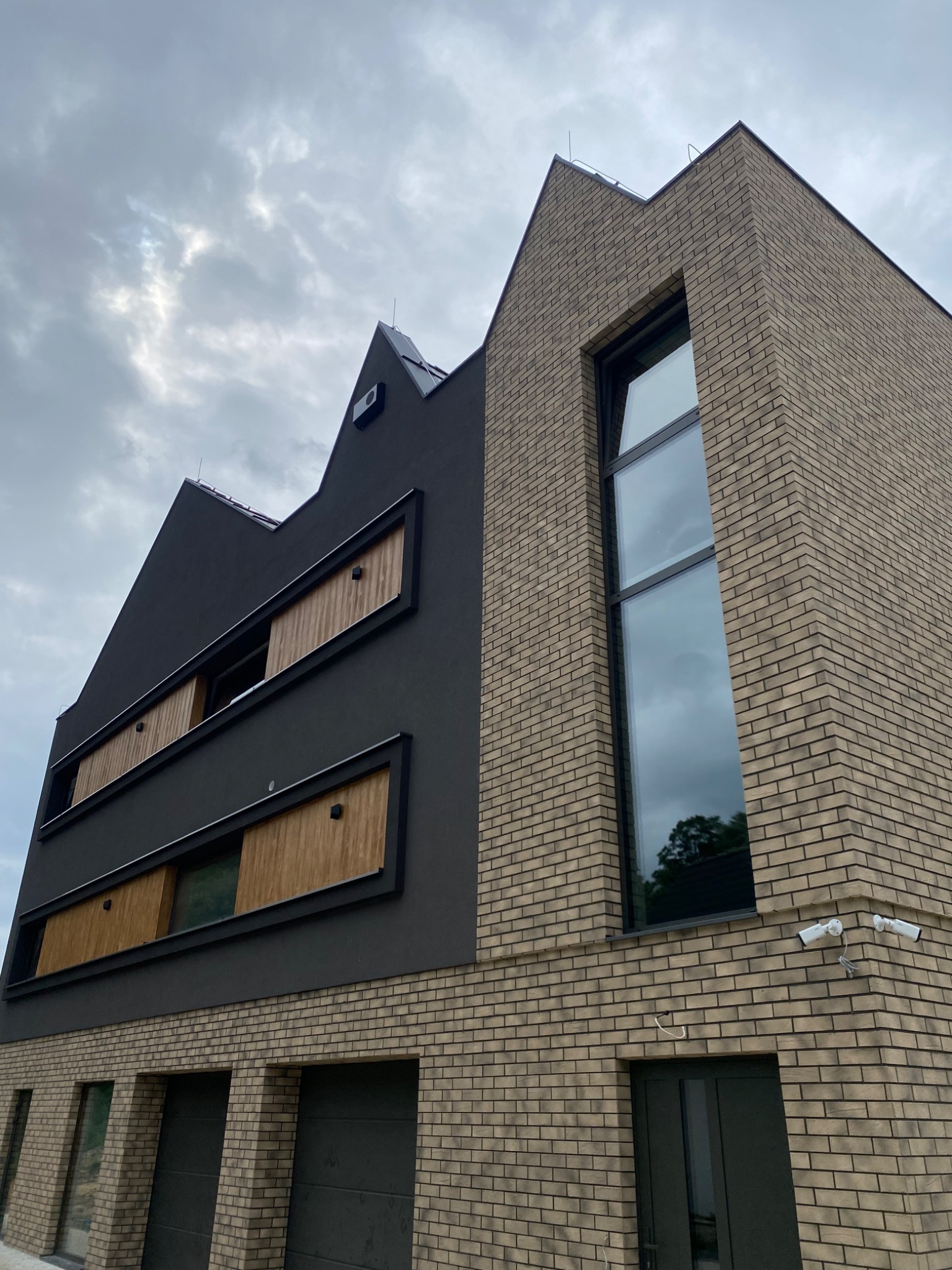 Nowoczesna elewacja budynku z cegły klinkierowej i szarego tynku, z drewnianymi elementami przesłon okiennych, widoczne kamery monitoringu i brama garażowa