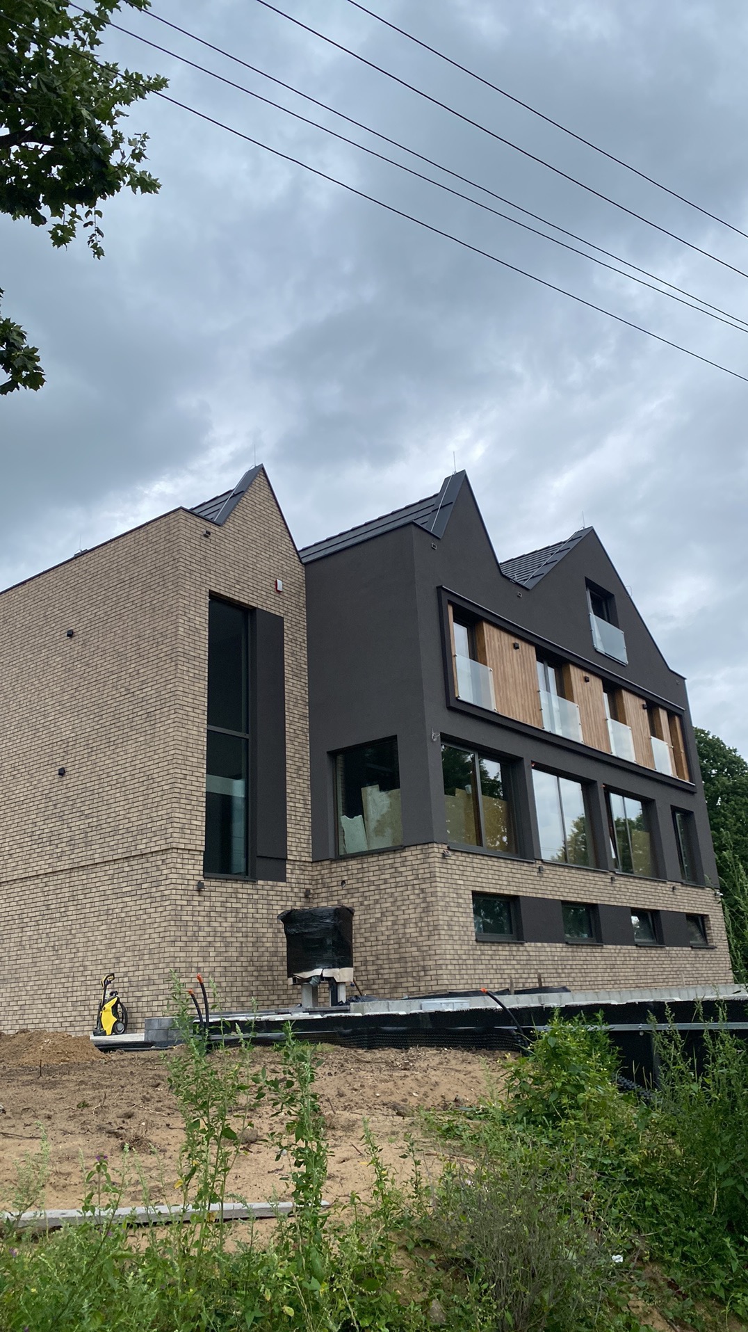 Nowoczesny dom dwukondygnacyjny z elewacją z cegły klinkierowej i fragmentami w ciemnym tynku, widoczne prace wykończeniowe wokół budynku, żółta myjka ciśnieniowa na chodniku.