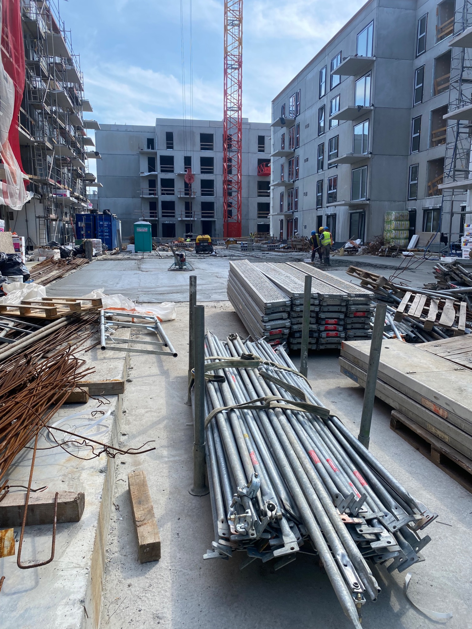 Budowa wielorodzinnego budynku z surową elewacją, widoczne rusztowania, dźwig i materiały budowlane, dwóch robotników w żółtych kamizelkach i kaskach.