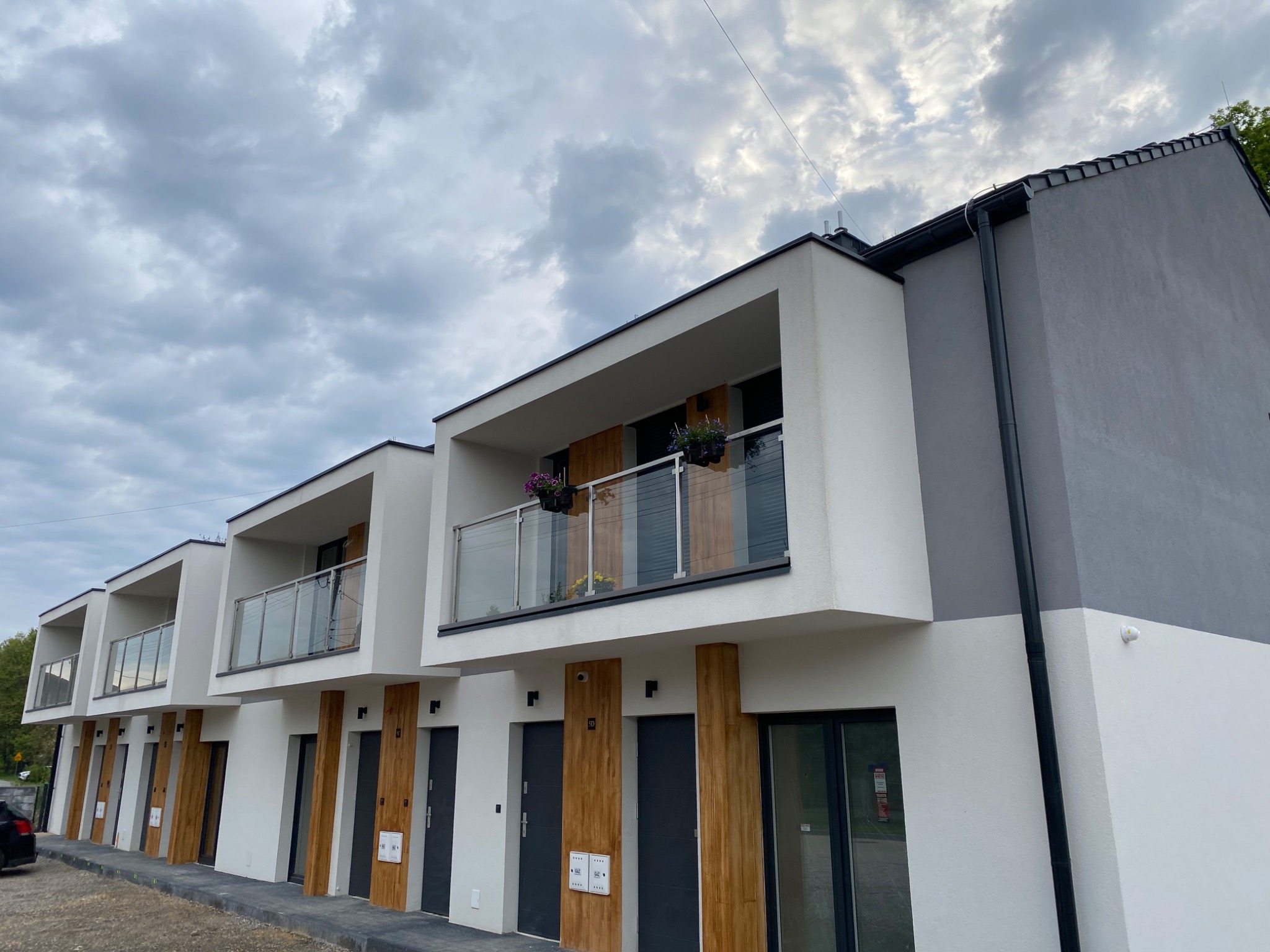Nowoczesne, szeregowe budynki mieszkalne z balkonami, wykończone w bieli i szarości z akcentami drewnopodobnymi, pod pochmurnym niebem.