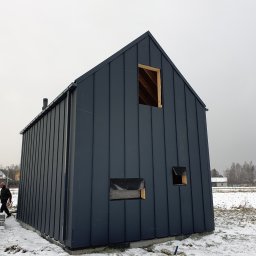 Mini Brda - Surowy, nowoczesny dom szkieletowy w trakcie budowy, z ciemną, metalową elewacją na tle zimowego krajobrazu, widoczne niezabezpieczone otwory okienne i fundament.