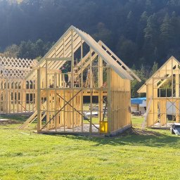 Mini Brda - Wznoszenie konstrukcji kilku domów z drewnianych belek na zielonej działce, widoczny pracownik na drabinie w jednym z budynków, las w tle.