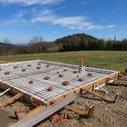 Mini Brda - Przygotowanie zbrojenia fundamentu pod budynek na nierównym terenie z widokiem na góry w tle, drewniany szalunek, cegły dystansowe i wystające rury kanalizacyjne.