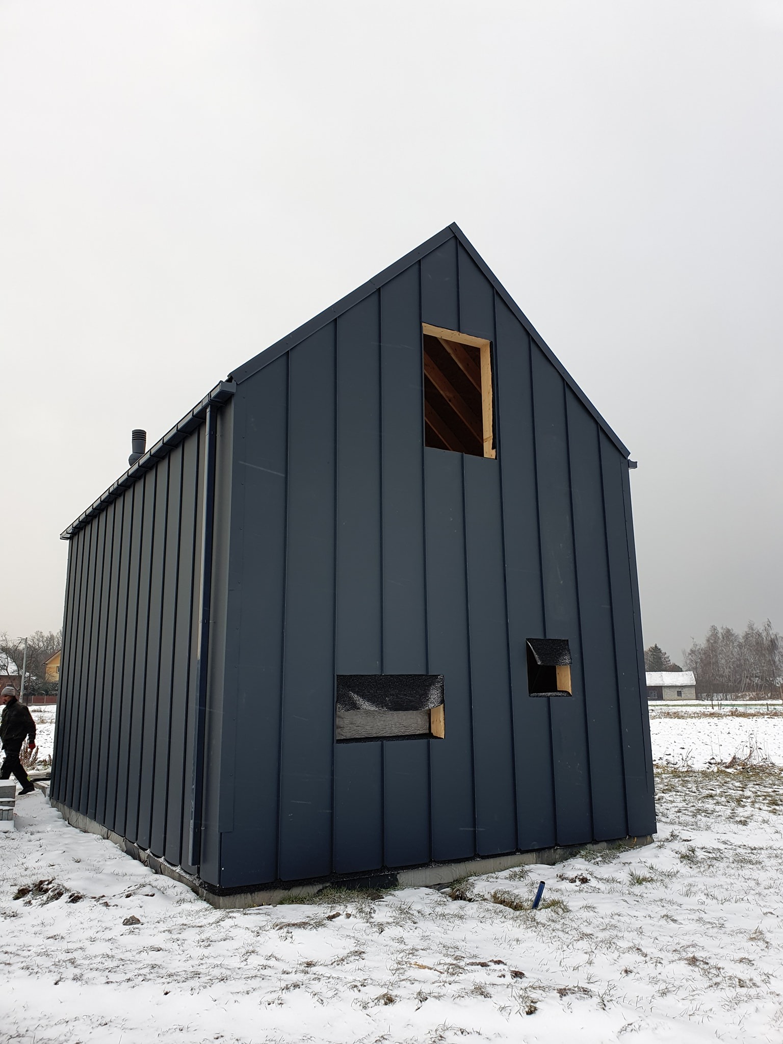 Surowy, nowoczesny dom szkieletowy w trakcie budowy, z ciemną, metalową elewacją na tle zimowego krajobrazu, widoczne niezabezpieczone otwory okienne i fundament.