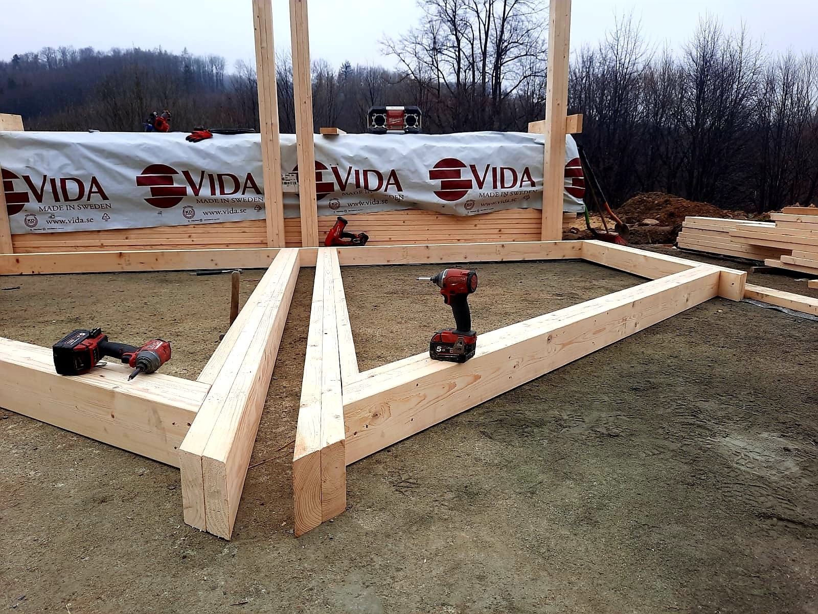 Wznoszenie drewnianej konstrukcji szkieletowej na fundamencie; widoczne narzędzia, deski z logo Vida i częściowo wzniesione ściany.