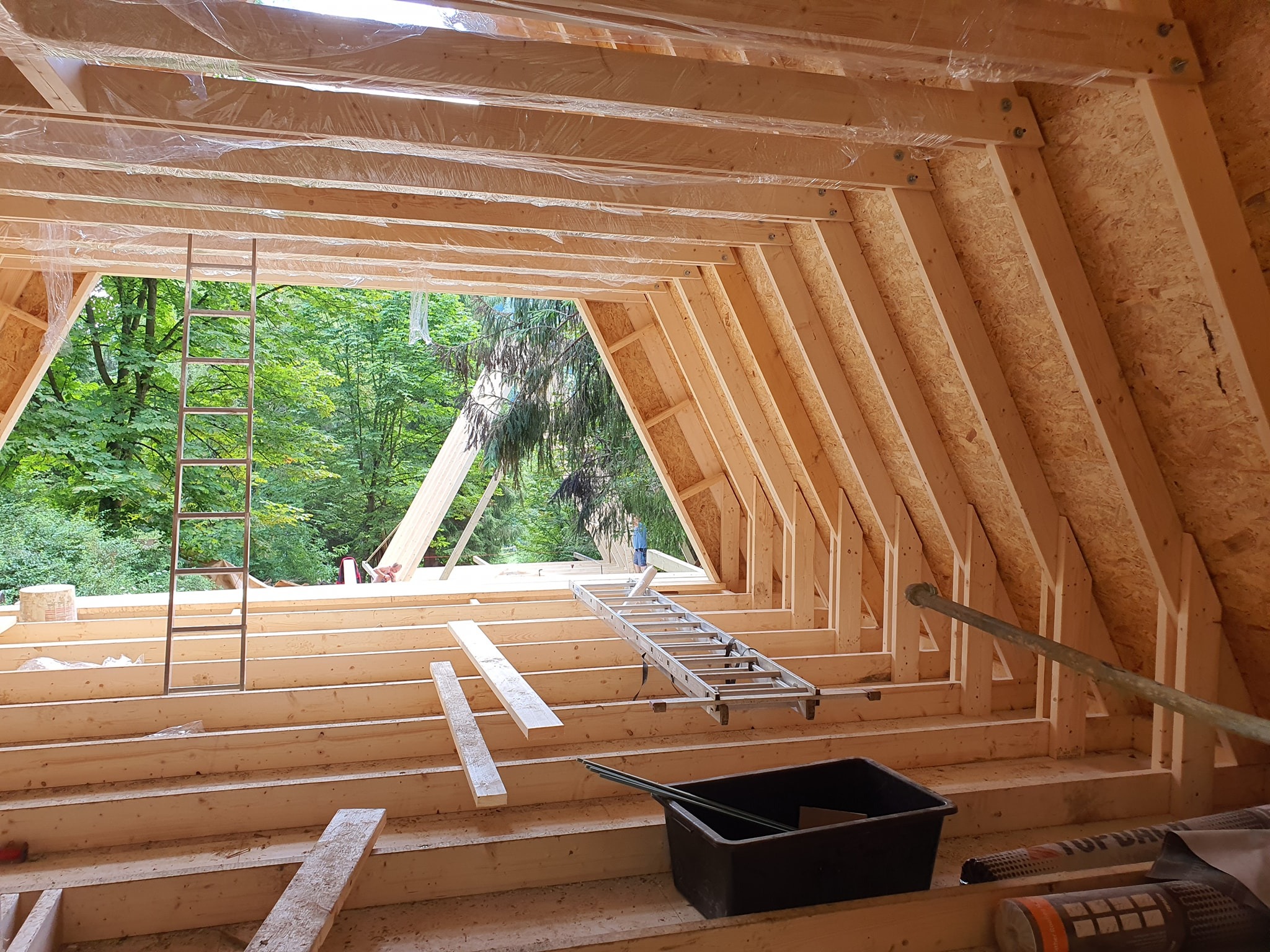Wnętrze domu w trakcie budowy: szkielet dachu z drewnianych belek, widoczne schody drabinkowe, narzędzia i folia ochronna, zieleń drzew w tle przez otwór w konstrukcji.