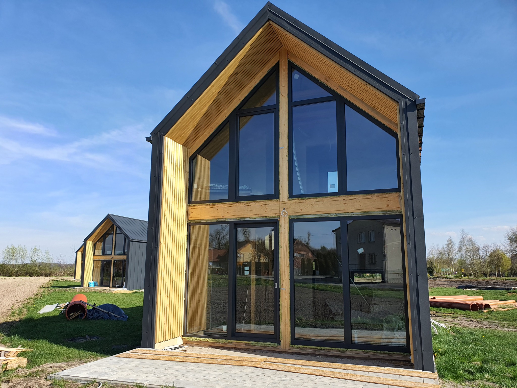 Nowoczesny dom z drewnianą elewacją i dużymi oknami, w trakcie budowy, widoczne elementy konstrukcyjne i przygotowane podłoże.