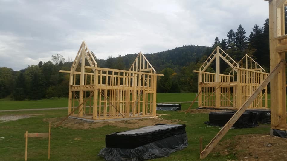 Wznoszenie konstrukcji z drewna na fundamencie podczas budowy domu z elementami szkieletowymi, widok na wzgórza w tle.