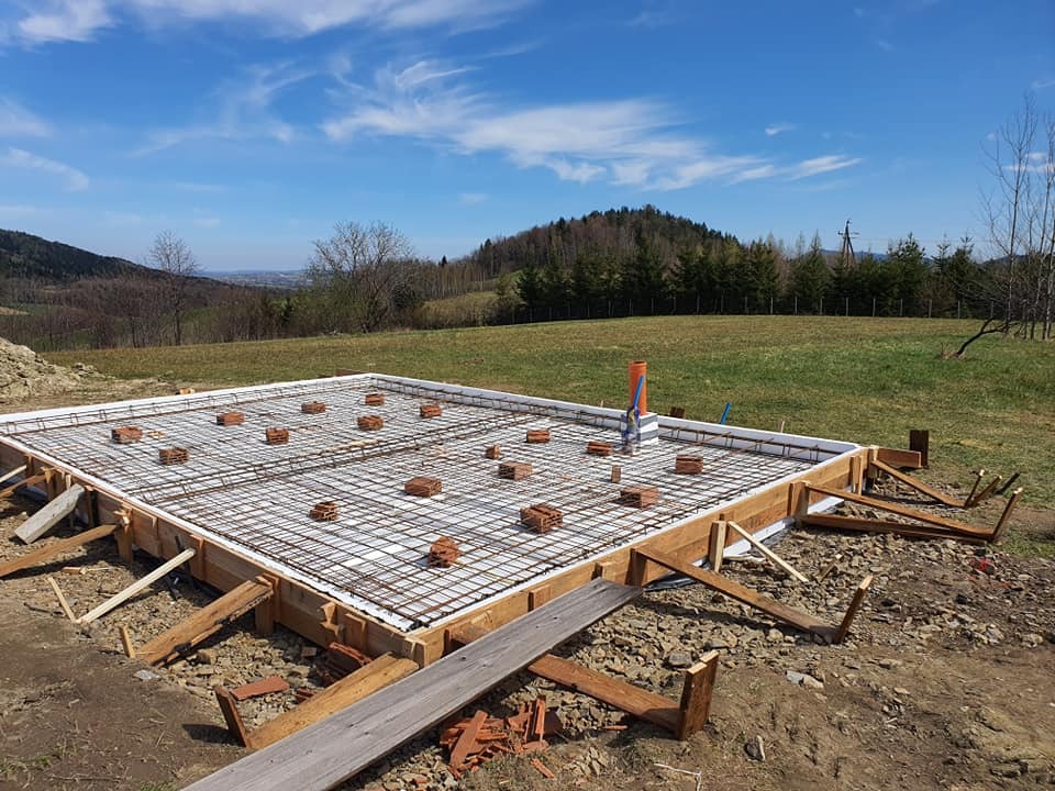 Przygotowanie zbrojenia fundamentu pod budynek na nierównym terenie z widokiem na góry w tle, drewniany szalunek, cegły dystansowe i wystające rury kanalizacyjne.