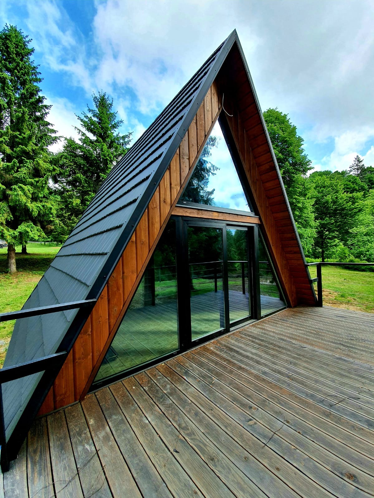 Nowoczesny dom w kształcie litery A z drewnianą elewacją i dużymi przeszkleniami, otoczony zielenią i drewnianym tarasem.