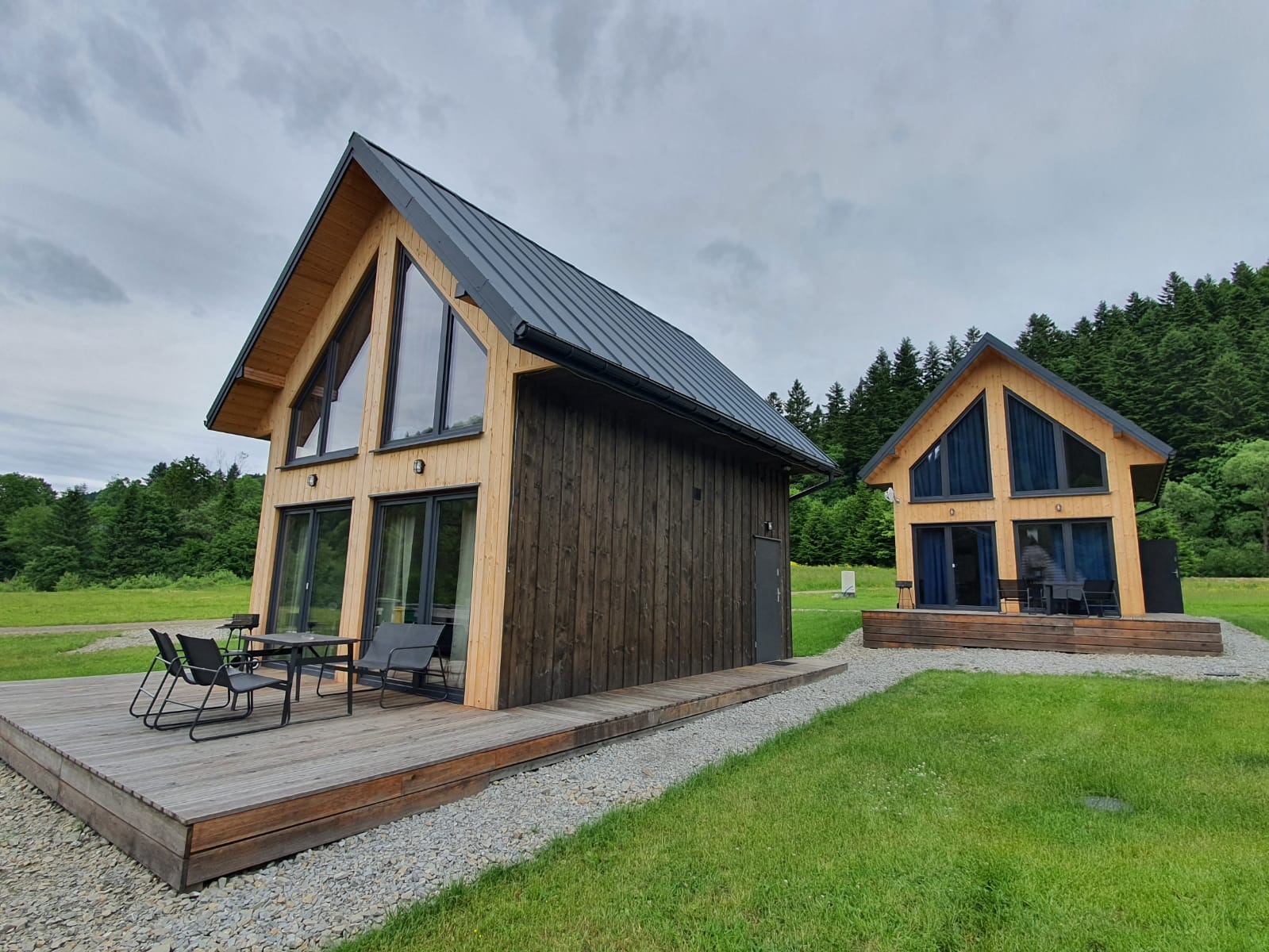 Dwa nowoczesne domki letniskowe z drewna, jeden z jasną elewacją i ciemnym pionowym panelem, drugi w tle z jasną elewacją, oba z dużymi przeszkleniami i ciemnymi dachami, otoczone zielonym...