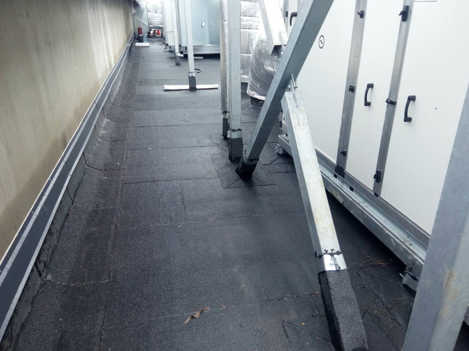 Czarna, płaska powierzchnia dachu pokryta papą termozgrzewalną, z widocznymi stalowymi konstrukcjami wsporczymi dla urządzeń wentylacyjnych i klimatyzacyjnych. Widoczne obróbki blacharskie przy...