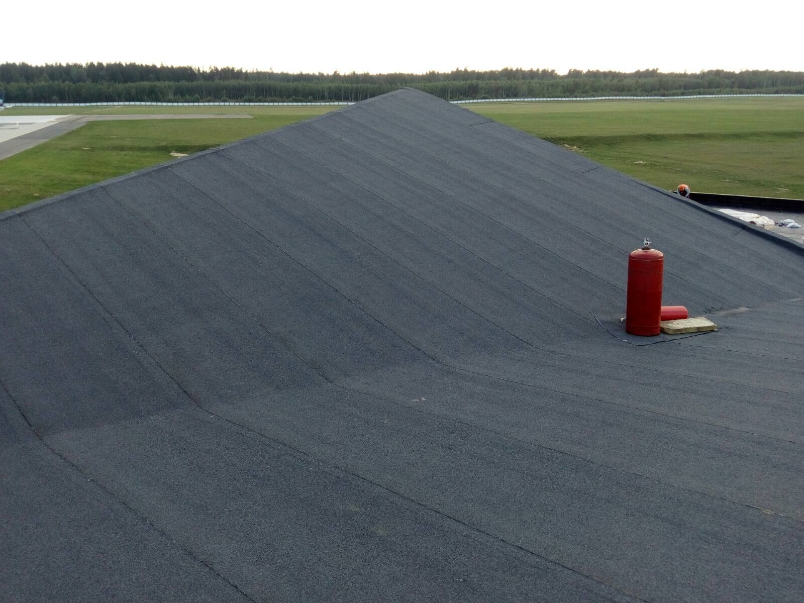 Wykończony dach pokryty papą termozgrzewalną z widocznym czerwonym zbiornikiem gazowym i elementami mocującymi, na tle zielonego pola i lasu.