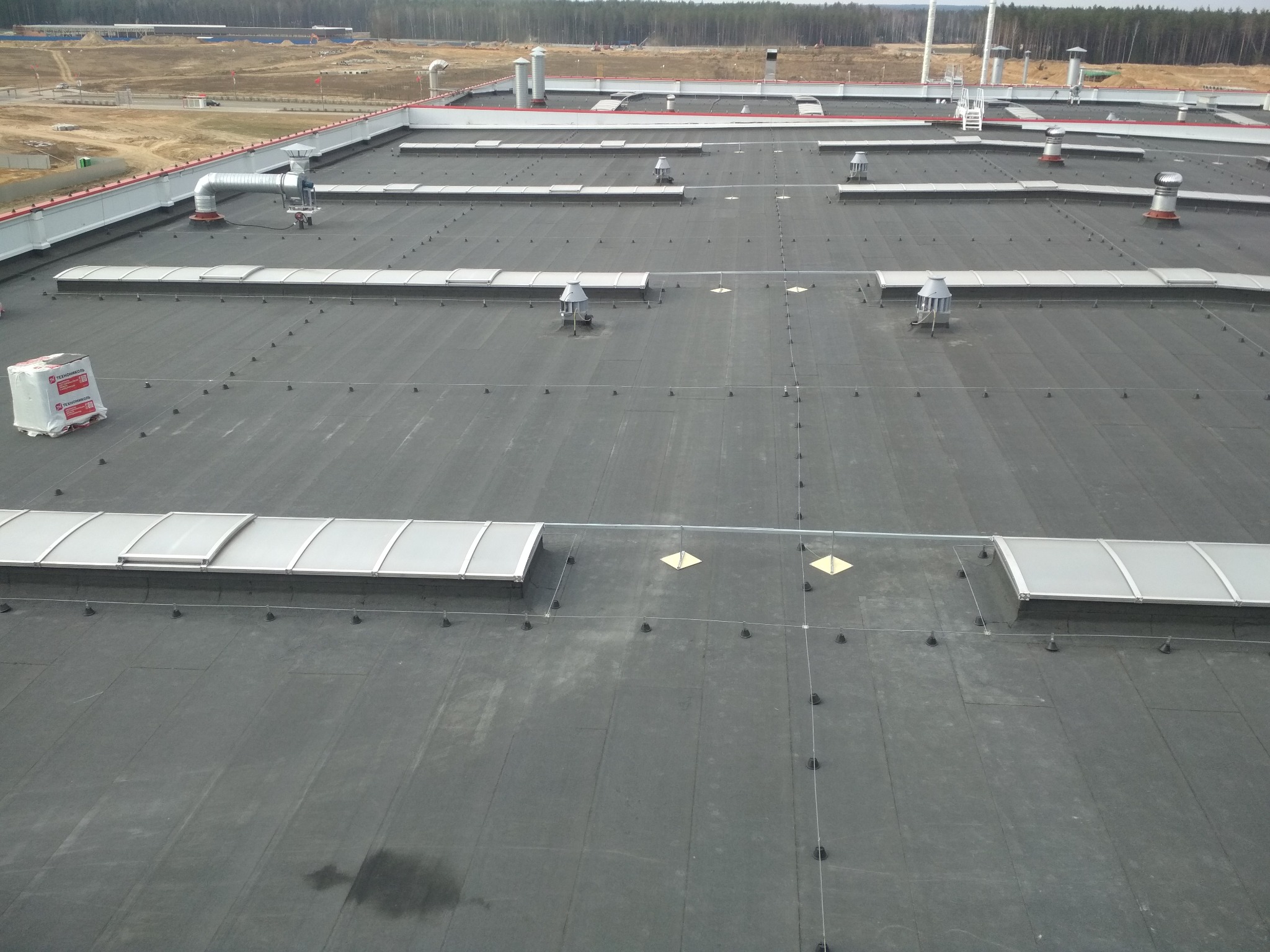 Rozległy, płaski dach pokryty czarną membraną bitumiczną z licznymi świetlikami, wywietrznikami i kominami wentylacyjnymi, widoczny z góry, z pakietem materiałów budowlanych w lewym górnym rogu.