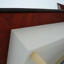 Fragment elewacji domu z drewnianą podbitką dachową w odcieniach brązu, białą ścianą i czarną rynną, sfotografowany z dołu.