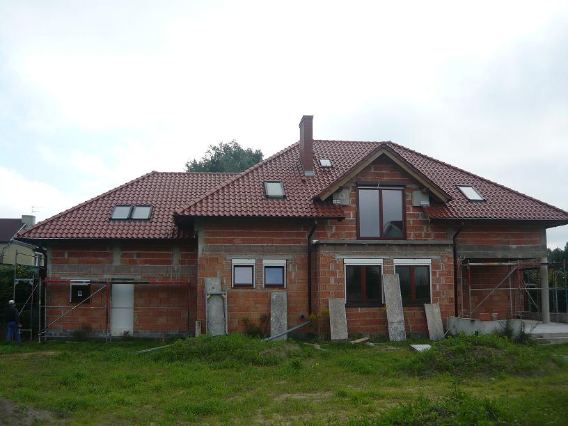 Niedokończony dom jednorodzinny z czerwonej cegły z zamontowanymi oknami i czerwoną dachówką, widoczne rusztowania i płyty betonowe na trawniku.