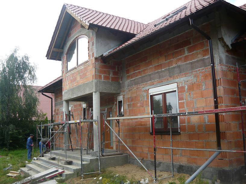 Budowa domu jednorodzinnego w stanie surowym otwartym: widoczne mury z czerwonej cegły, okna z roletami, częściowo wykonane schody wejściowe, rusztowania i pracujący robotnicy.