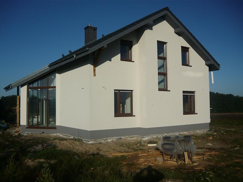 Nowoczesny dom jednorodzinny z białymi ścianami i ciemnobrązowymi oknami, widok z zewnątrz, z fragmentem ogrodu i niebem w tle. Widoczna dobudówka z dużymi przeszkleniami.