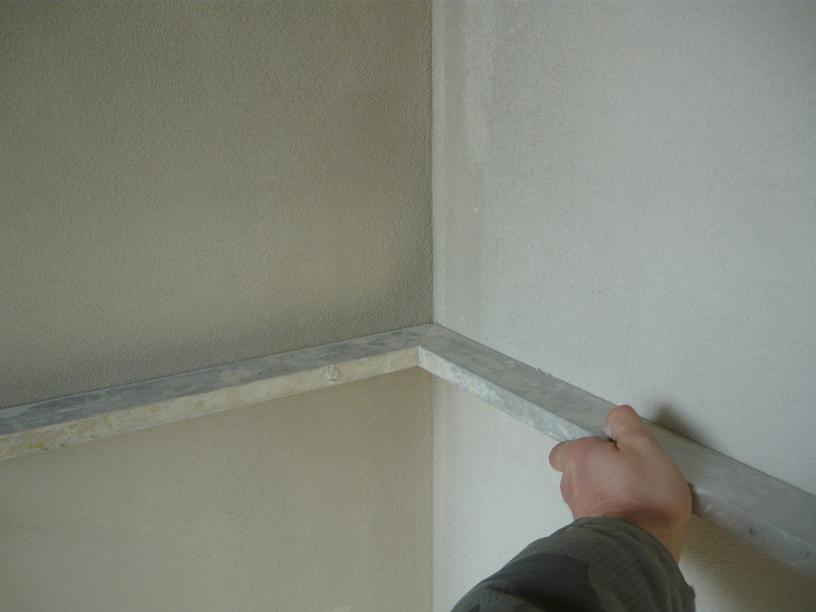 Przygotowanie narożnika ścian do tynkowania maszynowego, widoczny profil narożnikowy przytrzymywany przez dłoń, surowy stan wykończenia ścian.