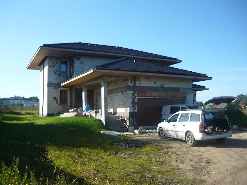 Niedokończony dom jednorodzinny w stanie surowym otwartym z ciemnym dachem, widoczna brama garażowa, zaparkowany biały samochód kombi z otwartym bagażnikiem i materiały budowlane na terenie budowy.