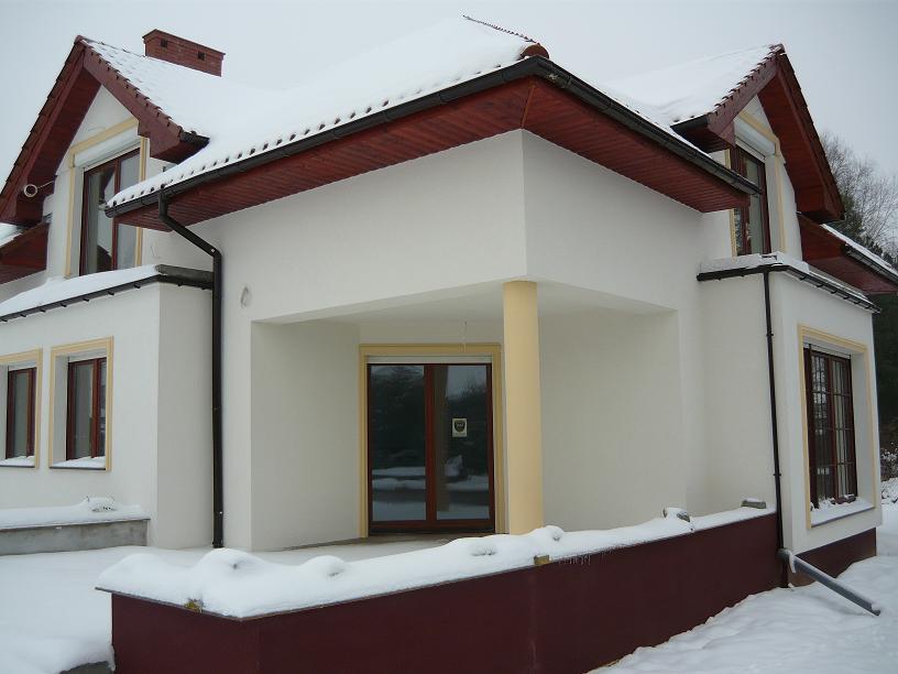 Wykończona elewacja domu jednorodzinnego w zimowej scenerii, z widocznymi oknami z roletami, rynnami i fragmentami ośnieżonego dachu.