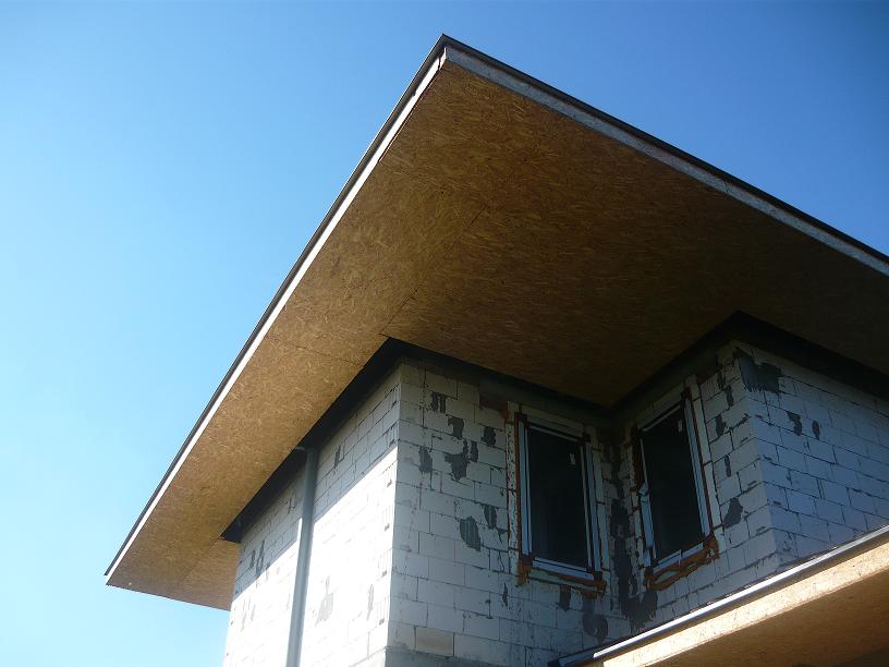 Surowa konstrukcja podbitki dachowej z płyty OSB na budynku w trakcie budowy, widoczne okna zabezpieczone folią i fragment ściany z betonu komórkowego.