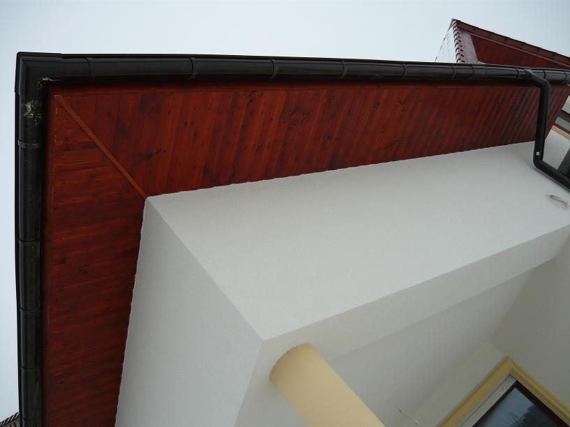 Fragment elewacji domu z drewnianą podbitką dachową w odcieniach brązu, białą ścianą i czarną rynną, sfotografowany z dołu.