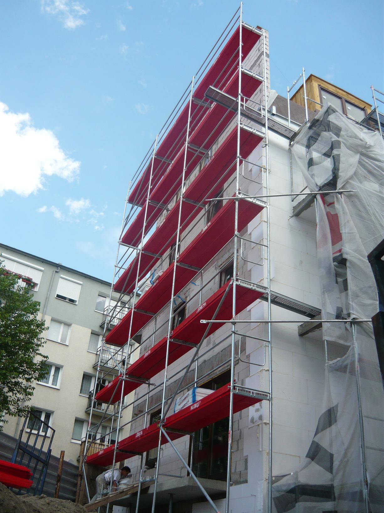 Budynek w trakcie ocieplania, obłożony rusztowaniami z czerwonymi podestami, widoczne białe płyty izolacyjne i pracownik na dole.