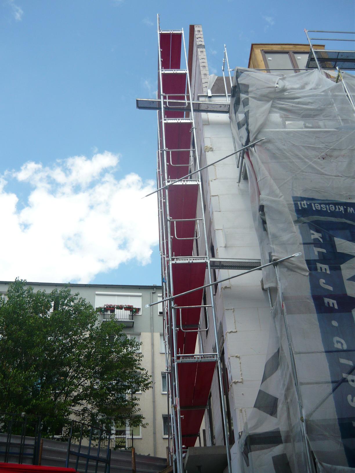 Budynek w trakcie ocieplania, widoczne rusztowanie z czerwonymi podestami, białe płyty izolacyjne i zabezpieczająca folia z nadrukiem, fragment sąsiedniego budynku z zielonym drzewem i błękitne...