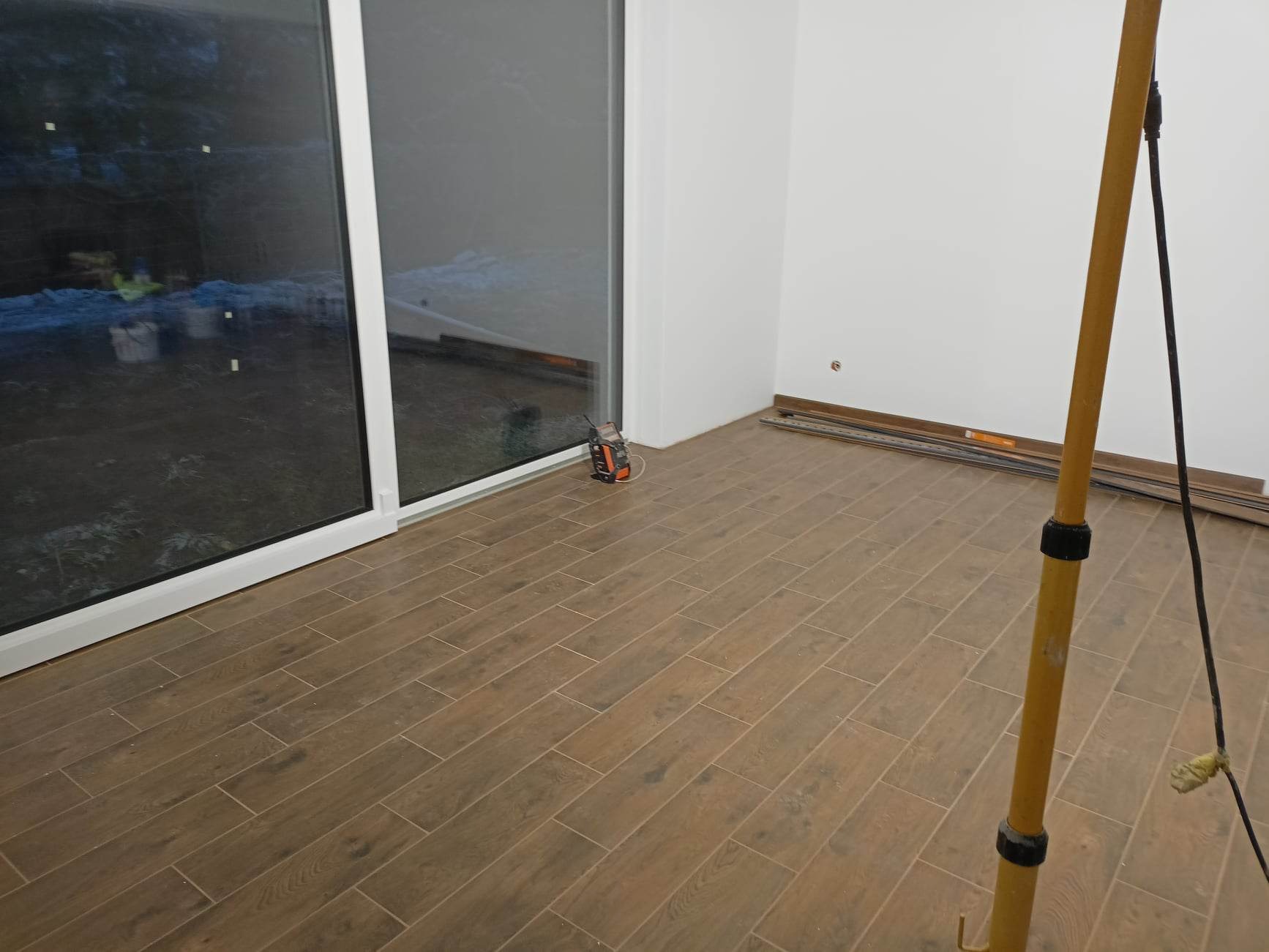 Wnętrze w trakcie remontu: brązowe płytki podłogowe imitujące drewno, białe ściany, przesuwne okno balkonowe, rozłożone listwy przypodłogowe i lampa budowlana.