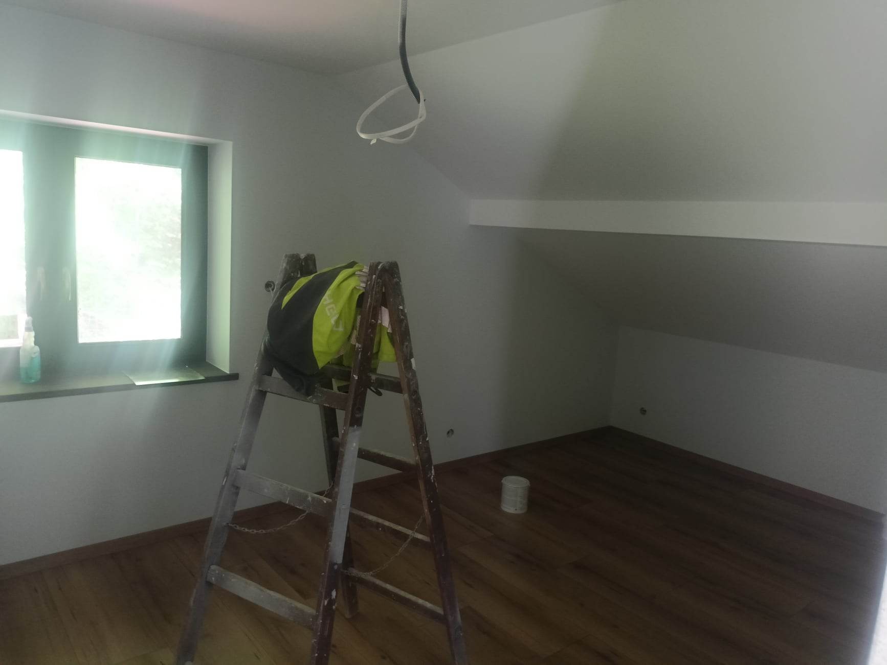 Pomalowane na biało poddasze z drewnianą podłogą, drabina z kurtką roboczą, puszka farby na podłodze, widoczne przewody elektryczne pod sufitem.