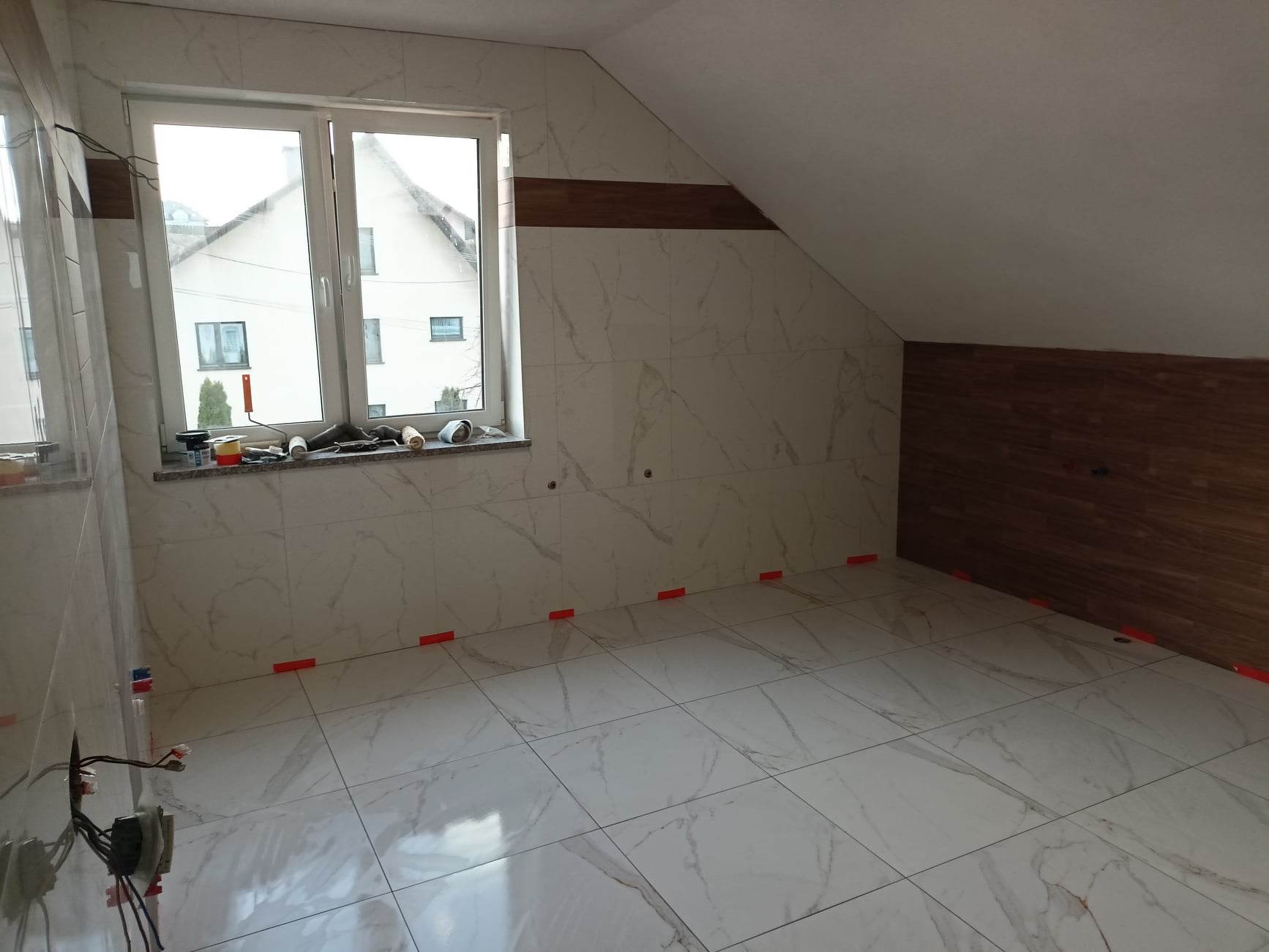 Płytki imitujące marmur na podłodze i ścianach poddasza, z widocznymi krzyżykami dystansowymi, narzędzia na parapecie okna, częściowo wykończona ściana z paneli drewnopodobnych.