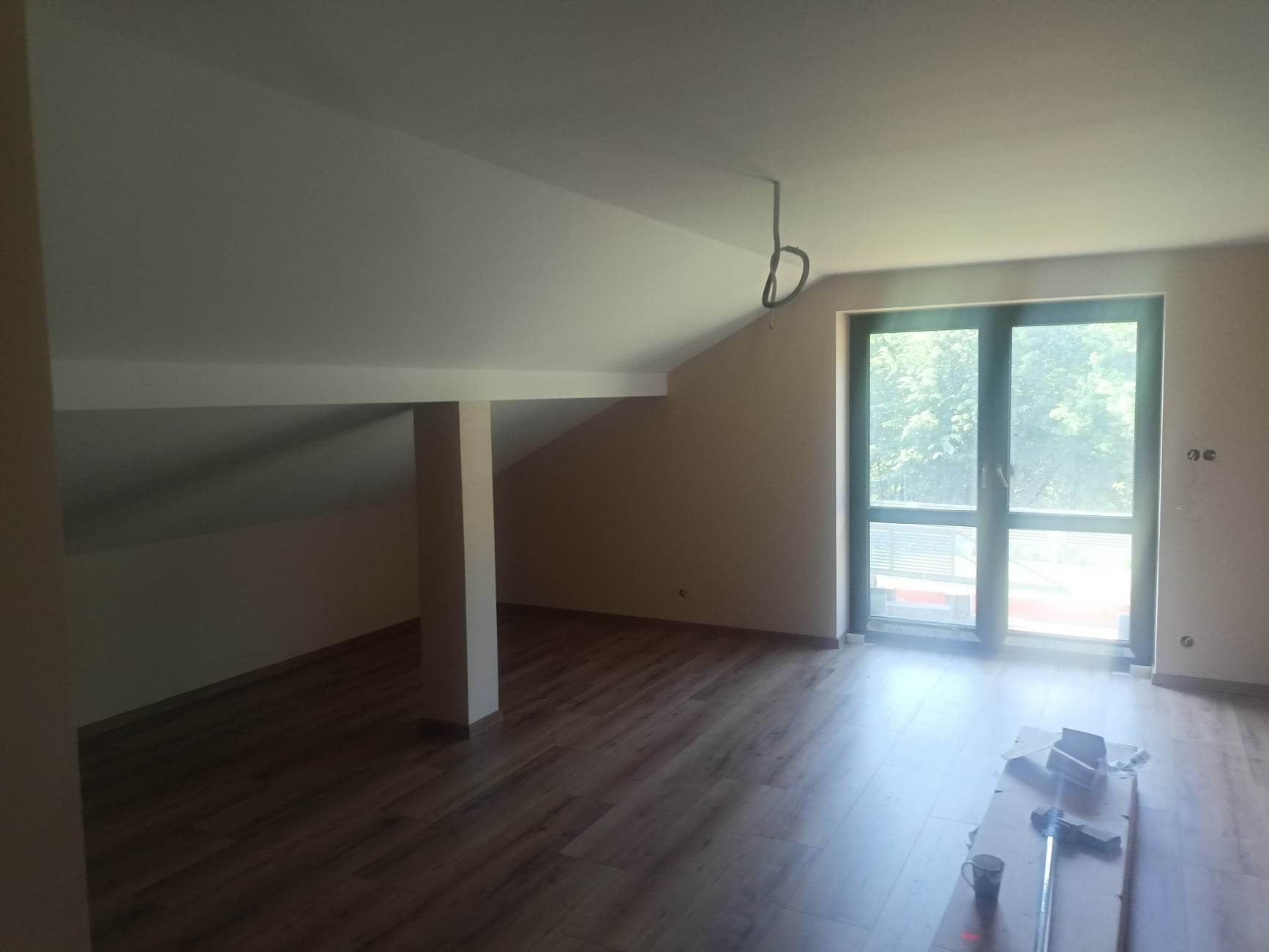 Poddasze po remoncie z drewnianą podłogą, świeżo pomalowanymi ścianami w neutralnym odcieniu i dużym oknem balkonowym, na podłodze kartonowe zabezpieczenie i narzędzia malarskie.