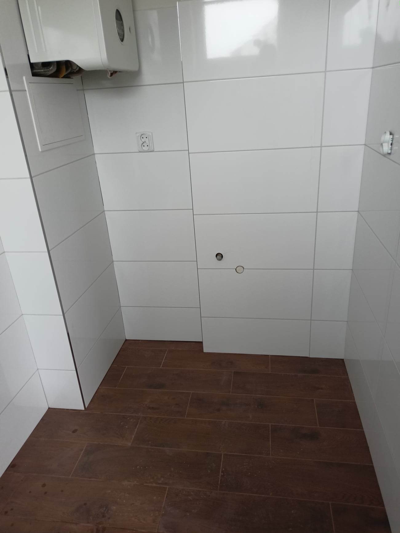 Nowoczesna łazienka z białymi, prostokątnymi płytkami na ścianach i podłogą wyłożoną brązowymi płytkami imitującymi drewno, widoczny bojler i gniazdko elektryczne.