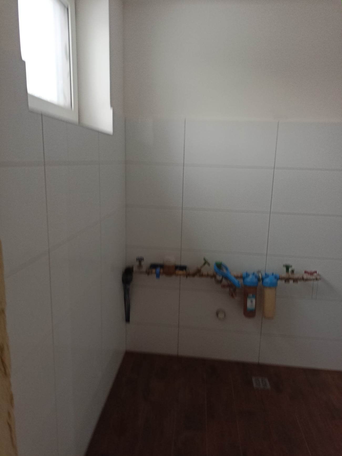 Białe płytki ścienne w łazience z zainstalowanym systemem filtracji wody na miedzianych rurach, widoczny fragment drewnopodobnej podłogi i okno u góry.
