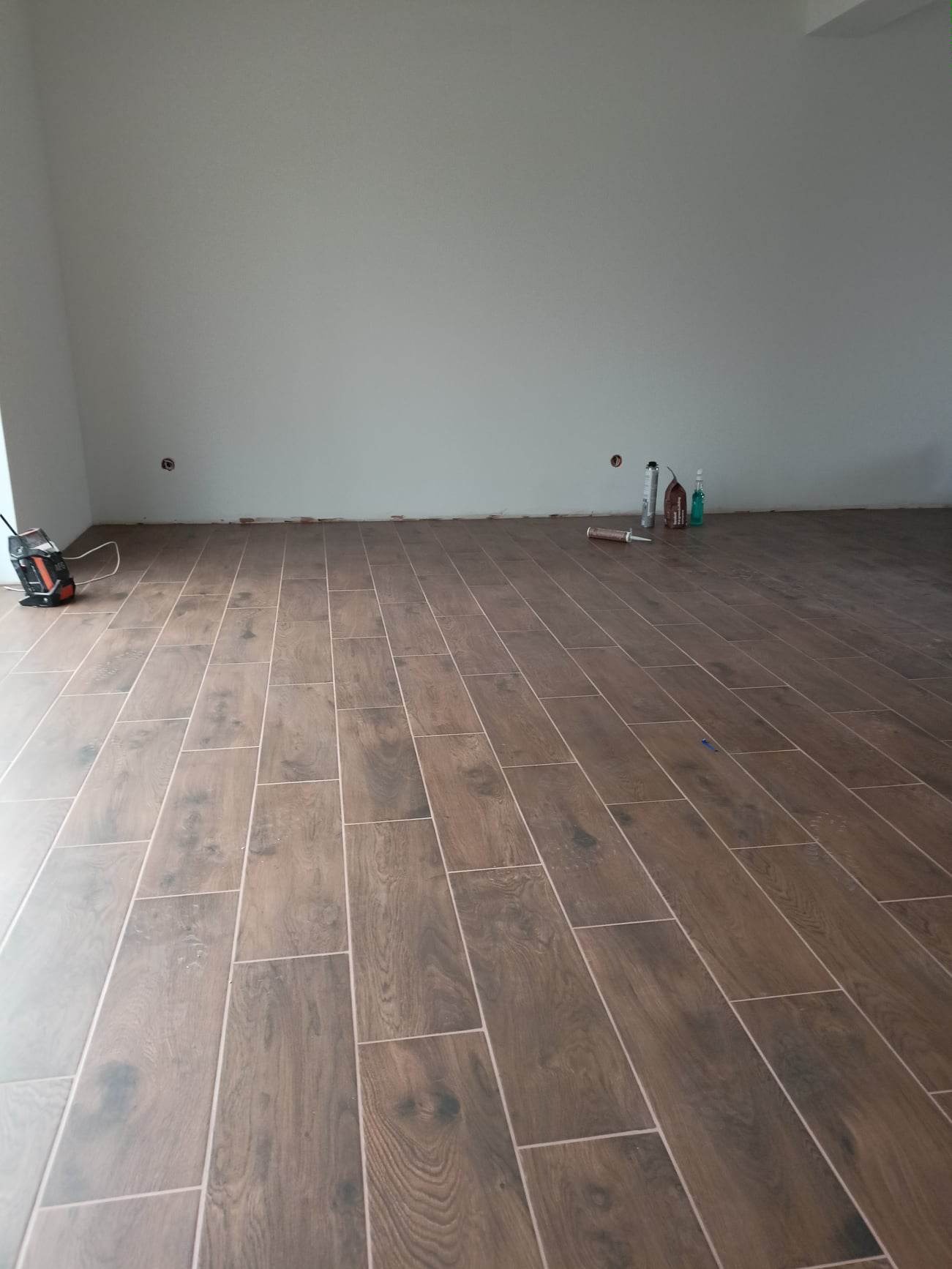 Ułożona podłoga z płytek imitujących drewno w pustym pomieszczeniu, widoczne narzędzia i materiały budowlane przy ścianie, przygotowane do dalszych prac wykończeniowych.