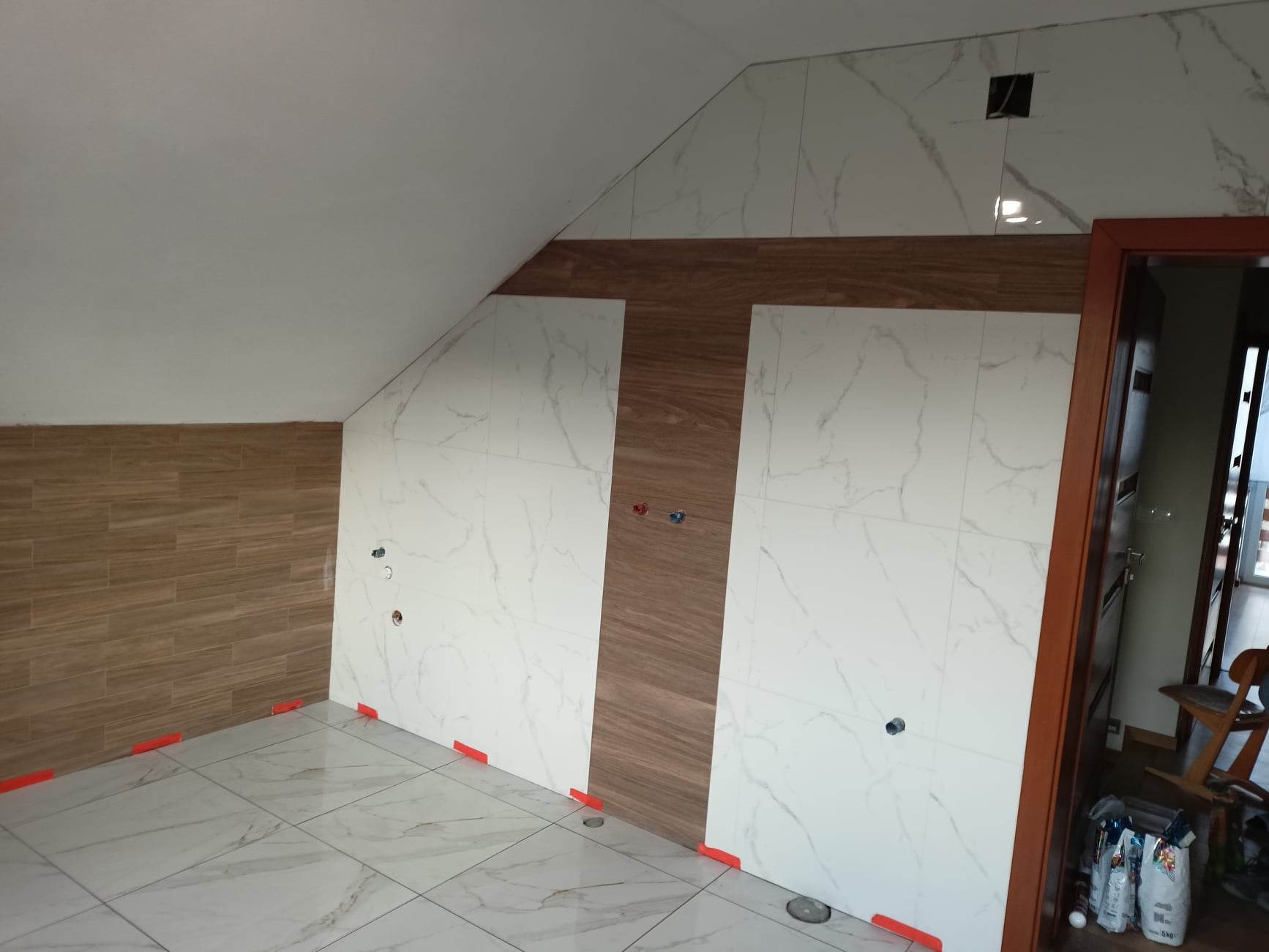 Łazienka w trakcie wykańczania, białe płytki imitujące marmur na ścianach i podłodze, brązowe płytki imitujące drewno, pomarańczowe kliny poziomujące płytki, widoczne otwory na instalacje.