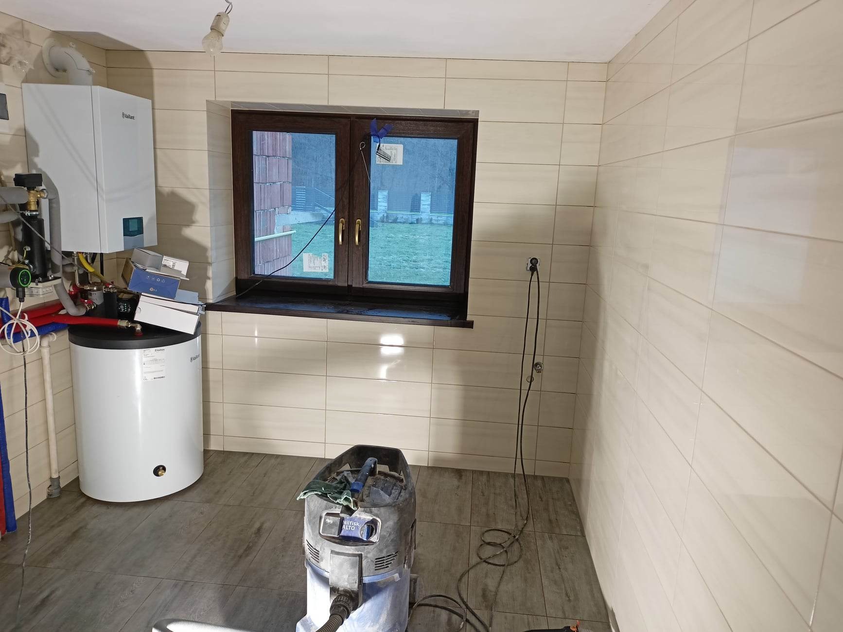 Wykończona łazienka z beżowymi płytkami, brązowe okno z widokiem na zewnątrz, biały bojler i odkurzacz przemysłowy na podłodze imitującej drewno.